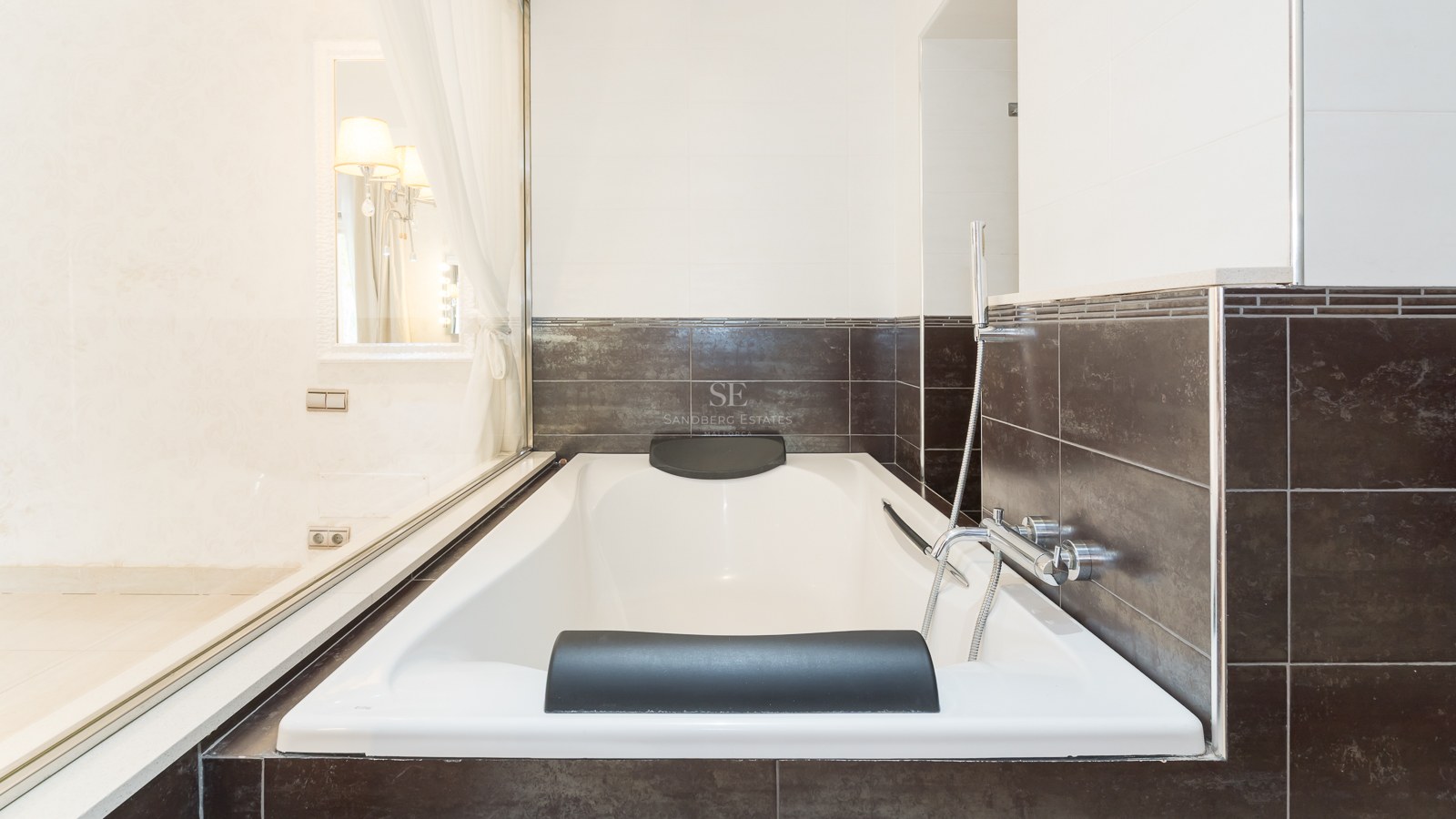 White built-in bathtub with black headrests and dark brown wall tiles in a modern bathroom.