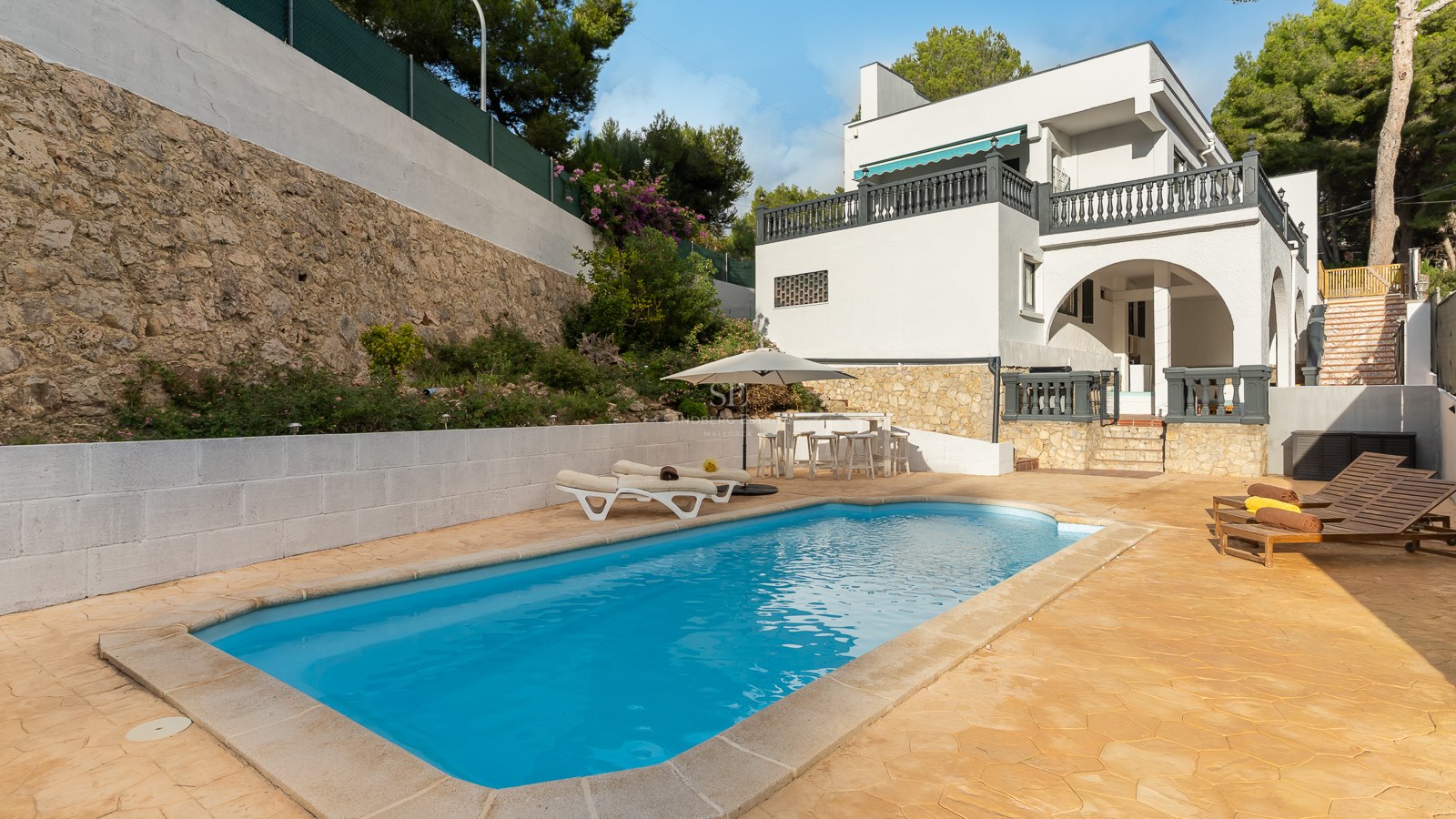 Rectangular swimming pool with turquoise water surrounded by a stone deck, sun loungers, and a white Mediterranean villa.