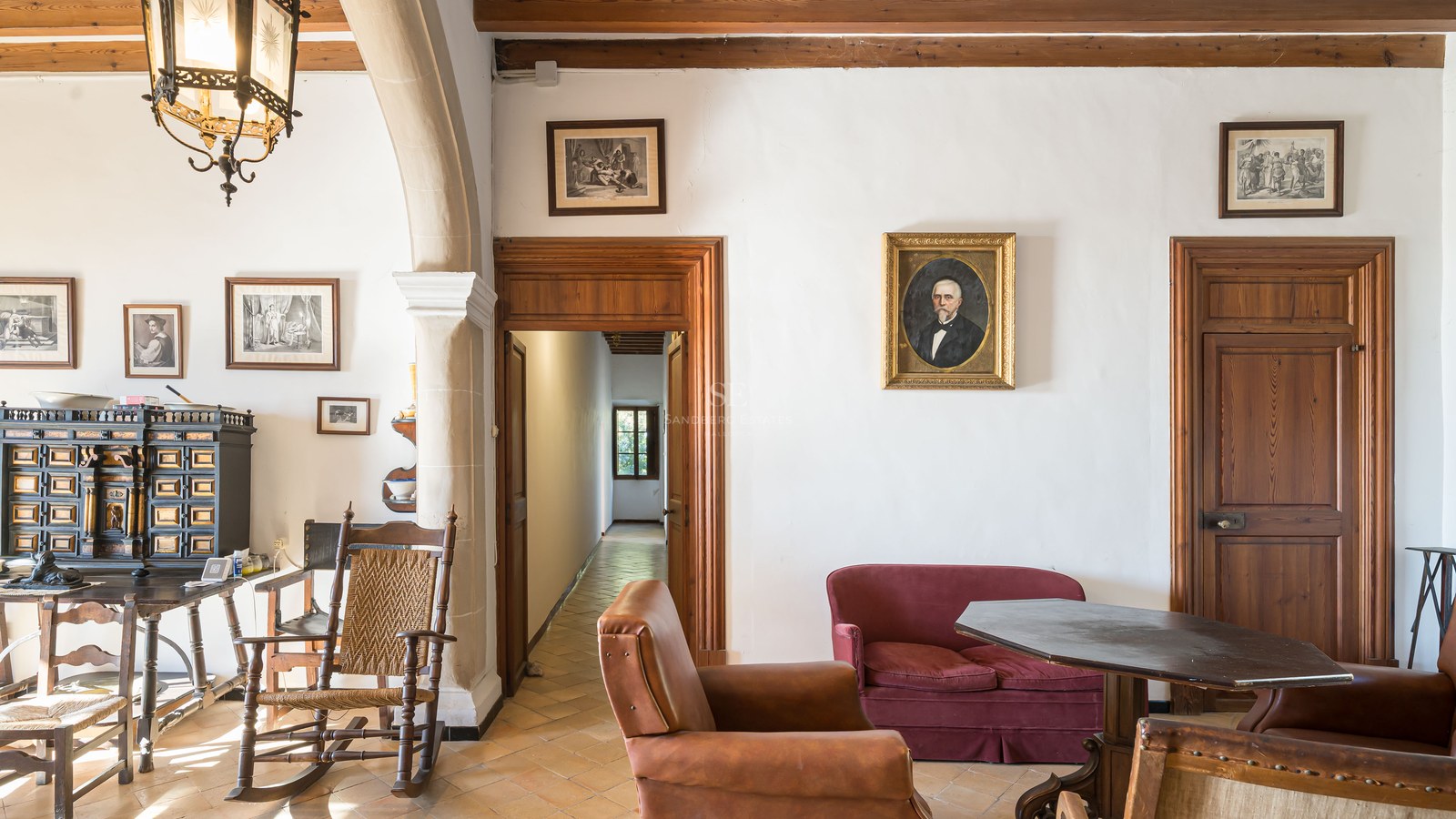 Salón clásico con muebles antiguos, suelos de terracota, sofá de terciopelo rojo y retratos en las paredes.