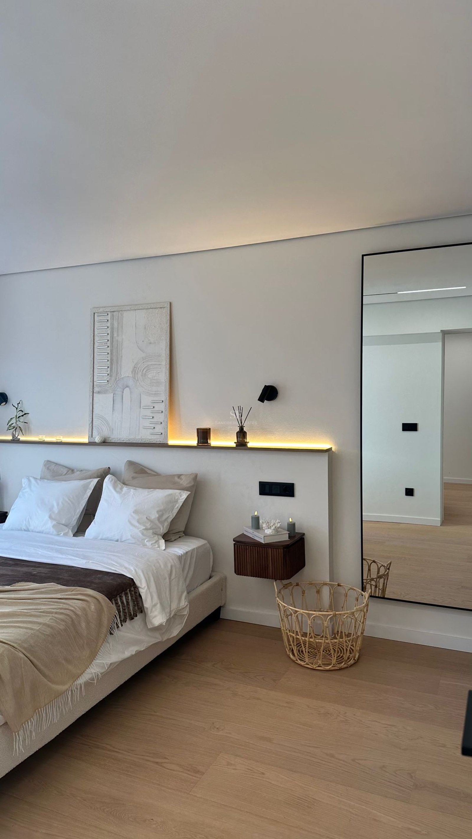 Chambre moderne avec lit blanc, rétroéclairage LED, œuvre d'art texturée et grand miroir sur parquet clair.