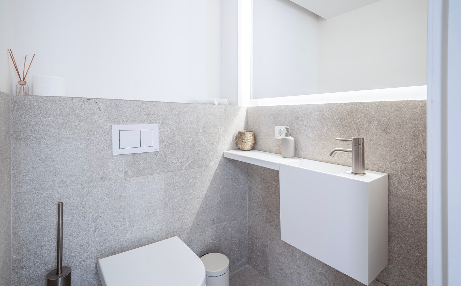 Minimalist bathroom featuring a white wall-hung toilet, small modern sink, and large backlit mirror against grey stone walls.
