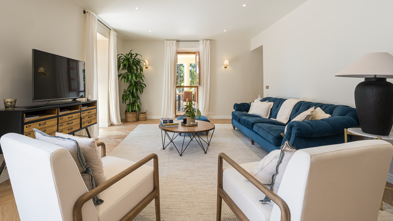 Bright living room featuring a navy velvet sofa, wooden armchairs, and chevron oak flooring leading to a terrace.