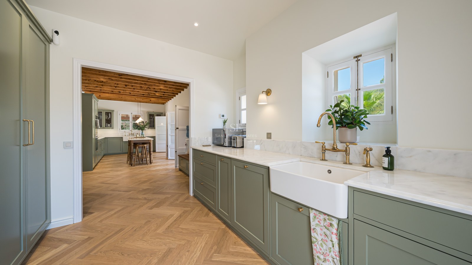 Sage green kitchen with marble countertops, brass taps, and herringbone wood flooring leading to a dining area.