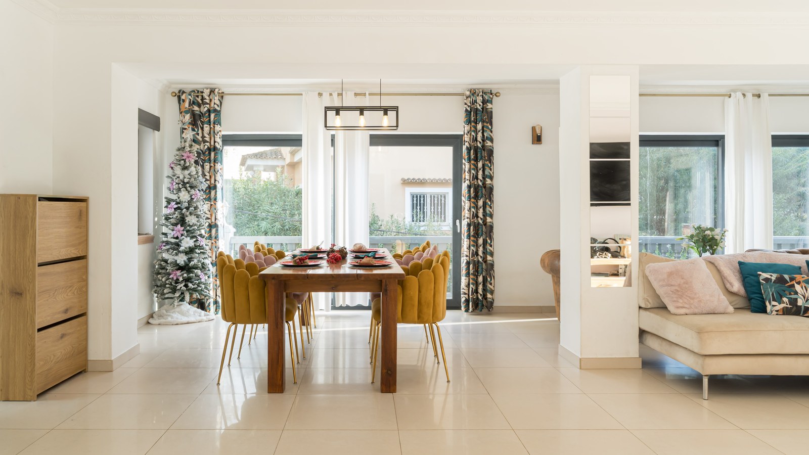 Modern dining area with a large wooden table, mustard velvet chairs, a Christmas tree, and large glass patio doors.