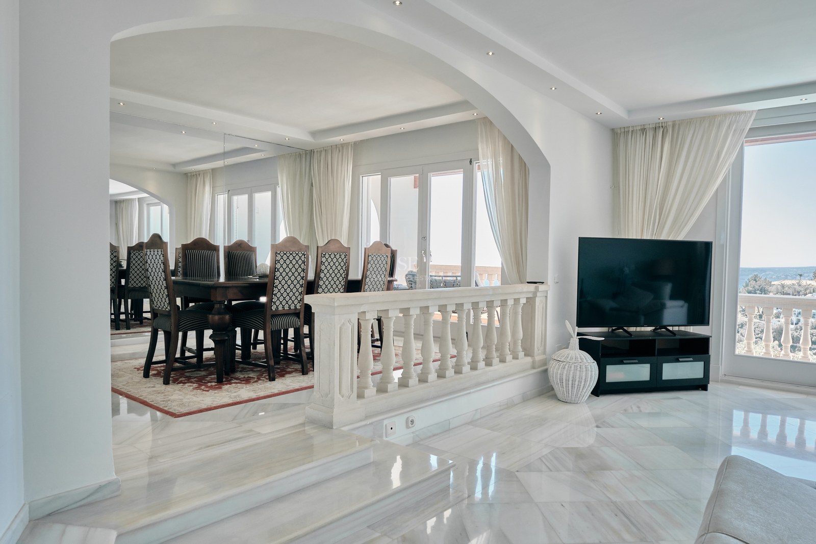 Bright split-level room with polished marble floors, an arched entrance to a dining area, and large terrace windows.