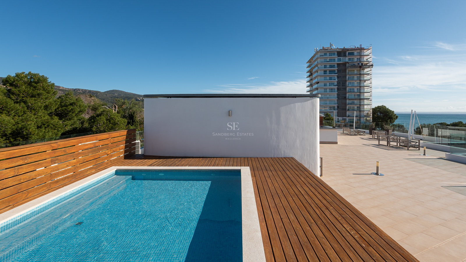 Modern rooftop swimming pool with wooden decking, overlooking the sea and mountains under a clear blue sky.