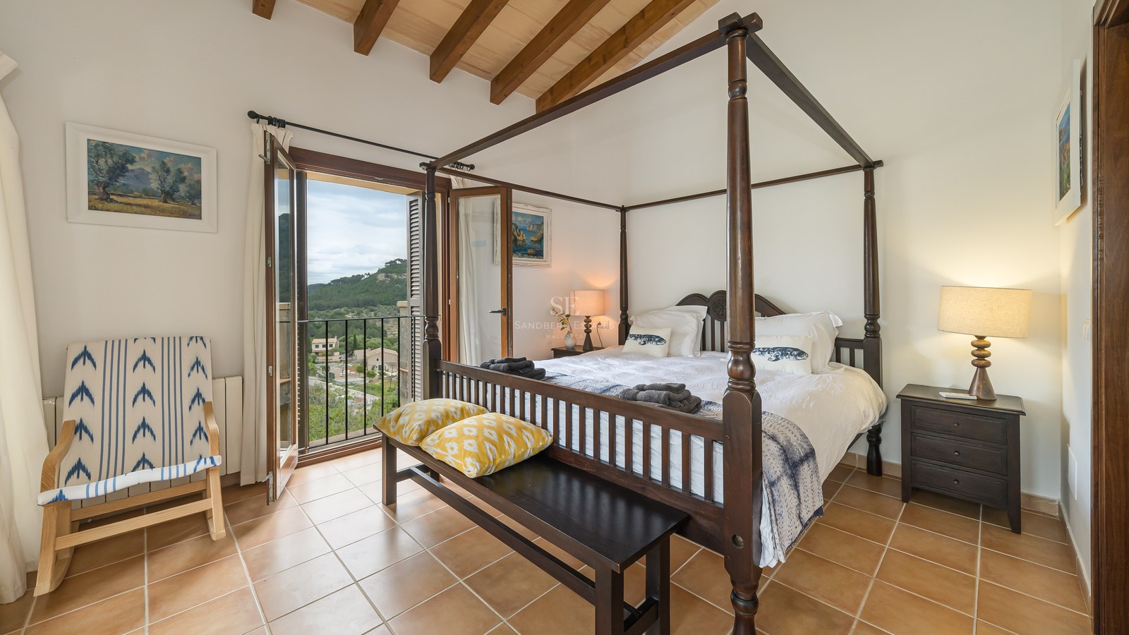 Master bedroom med himmelsäng, träbjälkar, terracottagolv och balkong med utsikt över bergen.
