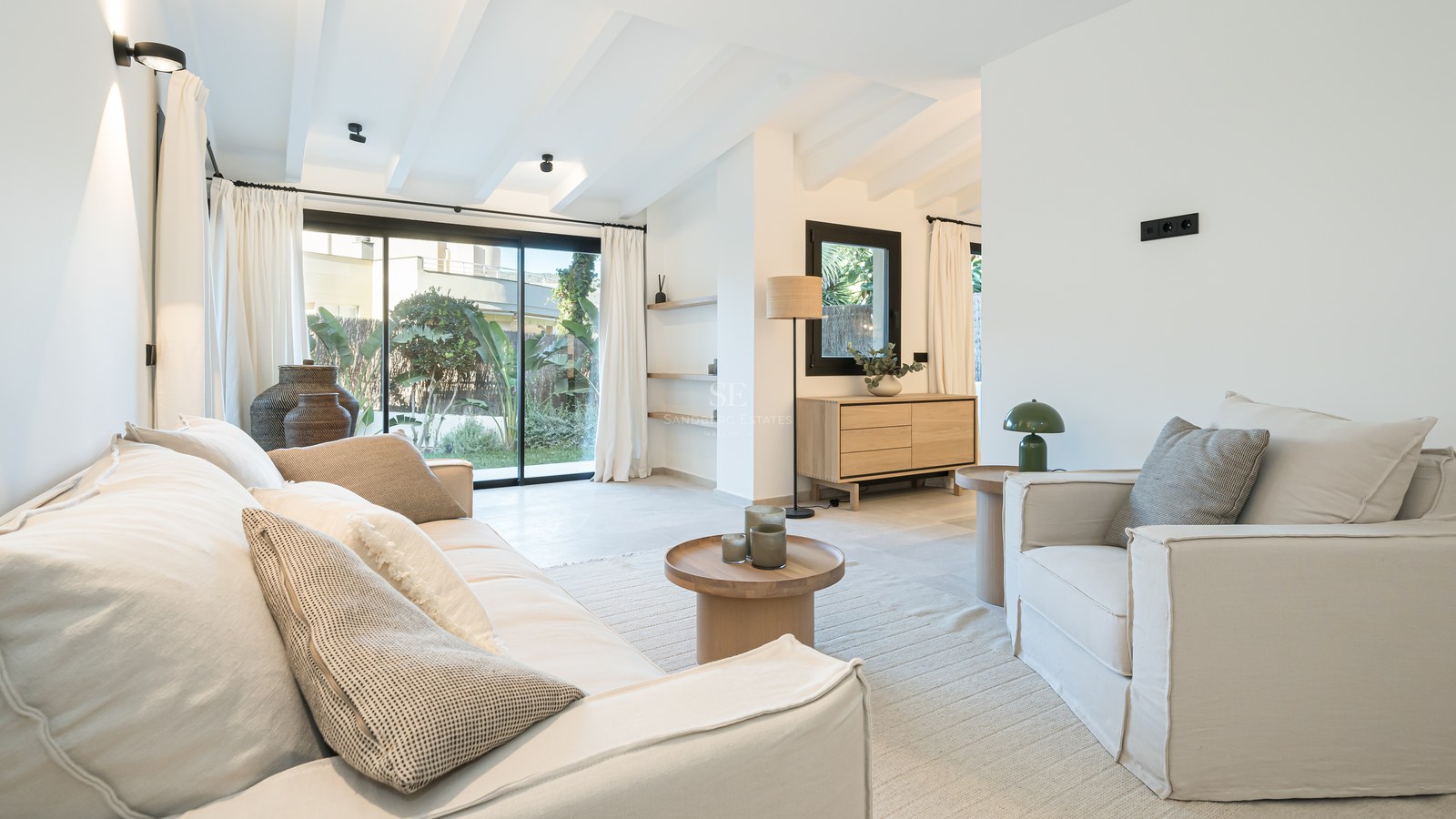 Salon lumineux avec plafonds à poutres blanches, canapés crème et baies vitrées ouvrant sur un jardin luxuriant.