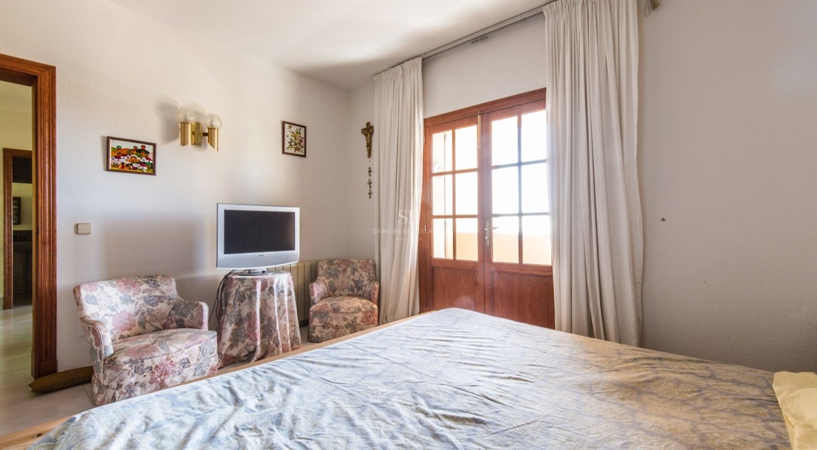 Chambre avec lit, deux fauteuils à fleurs et portes en bois ouvrant sur un balcon.