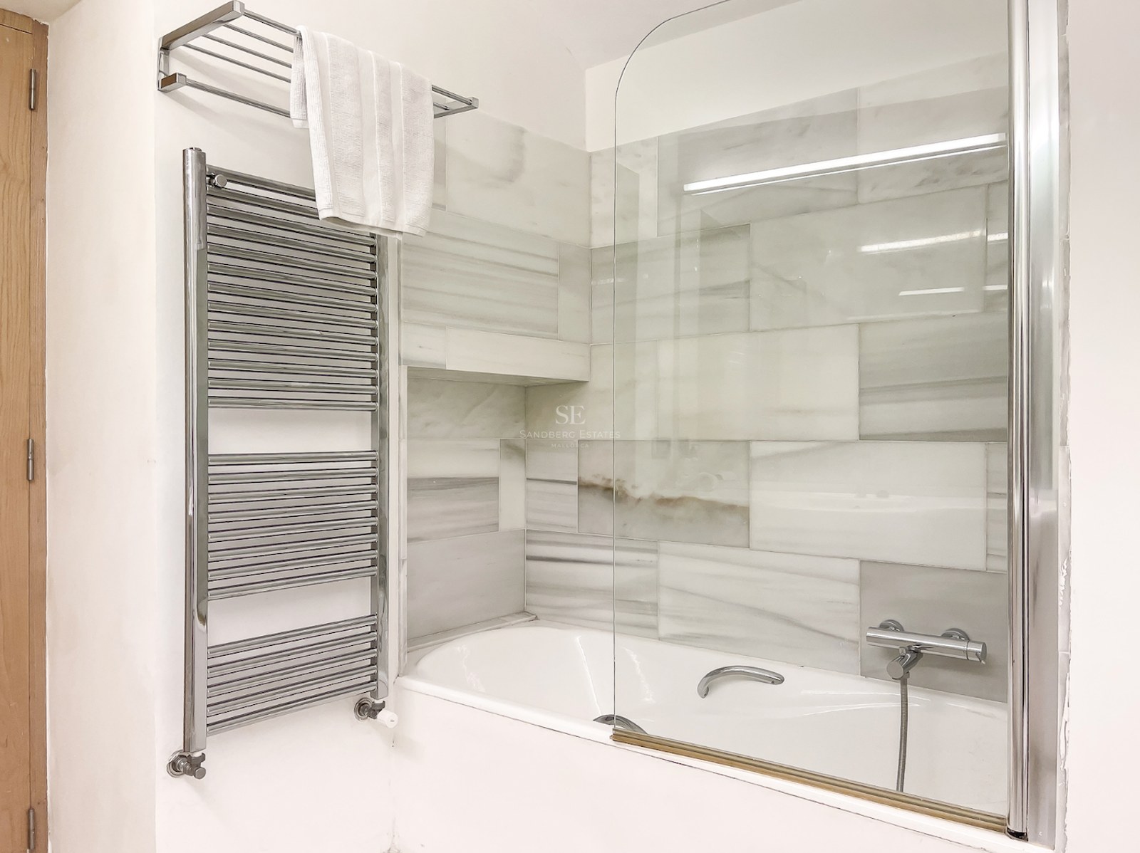 Bañera blanca con mampara de cristal, revestimiento de mármol gris y blanco y toallero radiador cromado.