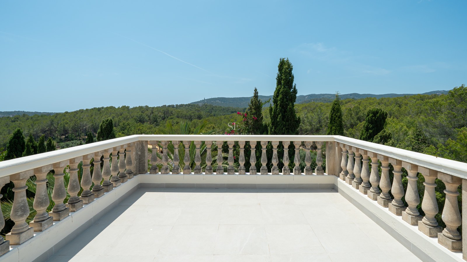 Spacious outdoor terrace with a classical stone balustrade overlooking a dense pine forest and hills under a clear blue sky.