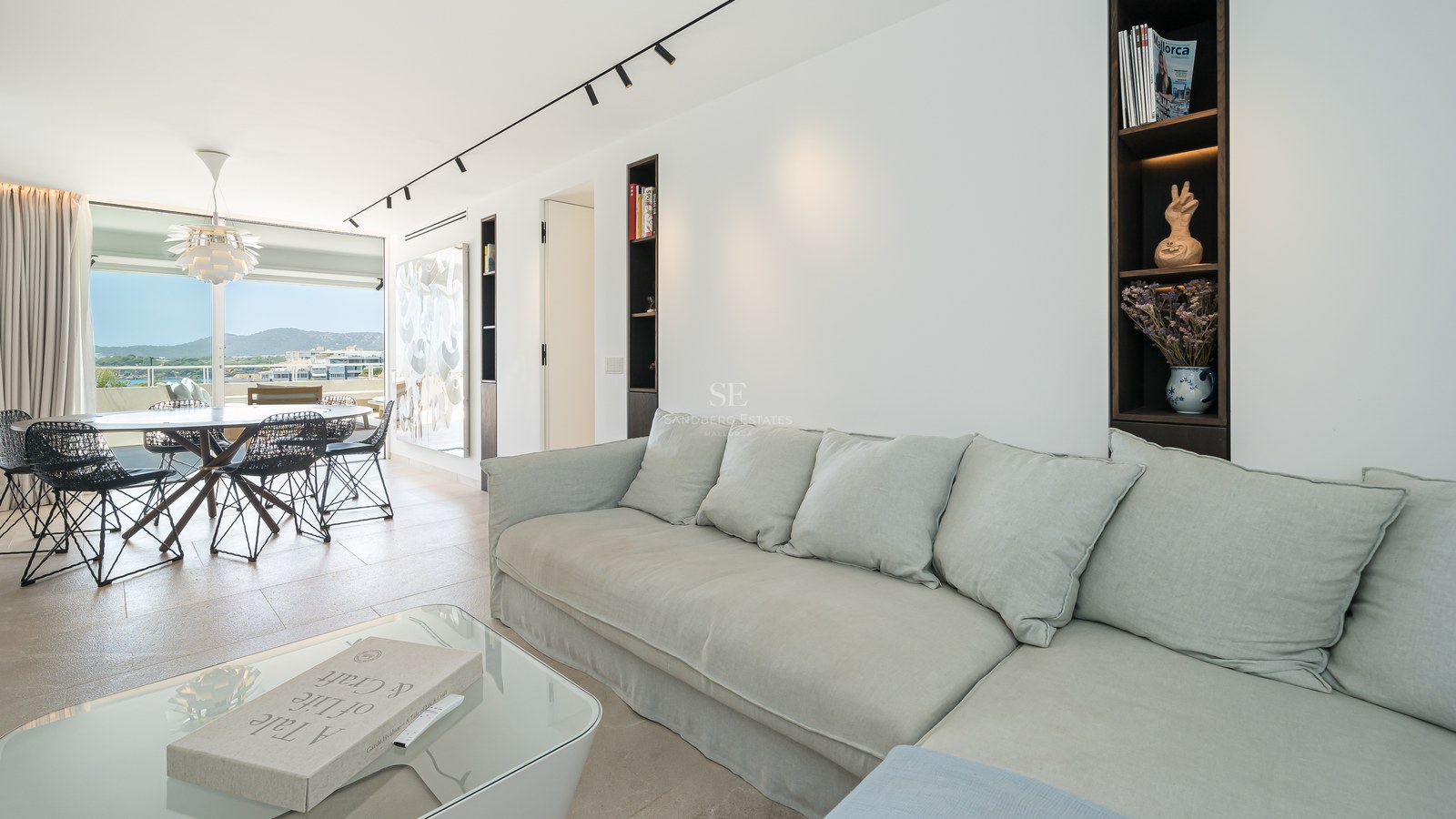 Salon lumineux avec un grand canapé gris clair, table basse en verre et salle à manger en arrière-plan avec vue sur la mer.