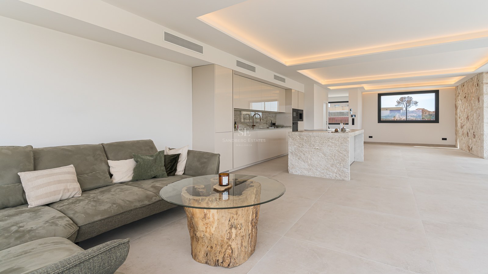Open-plan living room featuring an olive green sofa, a wood-trunk glass table, and a sleek minimalist white kitchen.