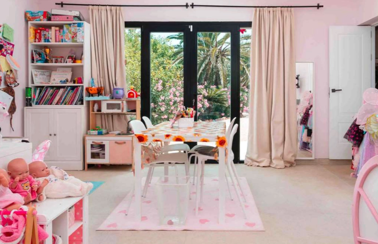 A sunlit children's playroom with pink walls and large glass doors opening to a Mediterranean garden with palms.