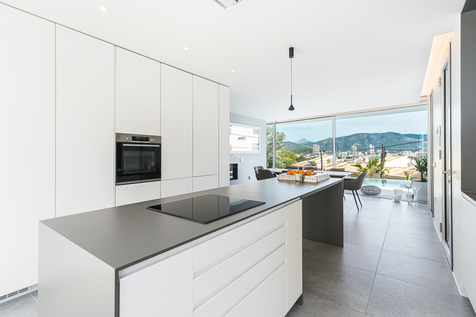 Cocina blanca luminosa con isla gris, placa de inducción y ventanales con vistas a las montañas y piscina.