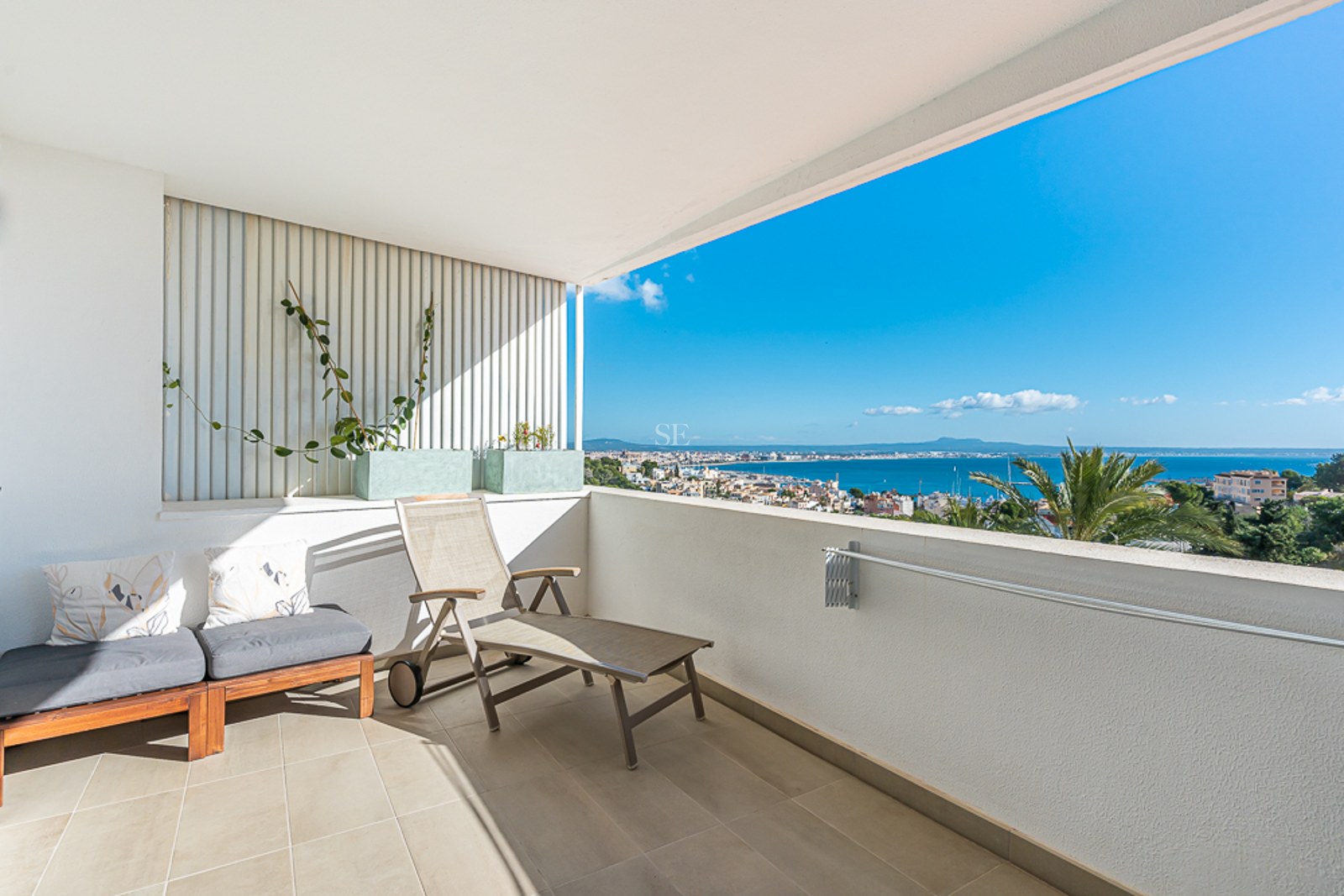Spacious balcony with glass railing, panoramic sea views, and modern relaxation furniture. Plants add a natural touch.