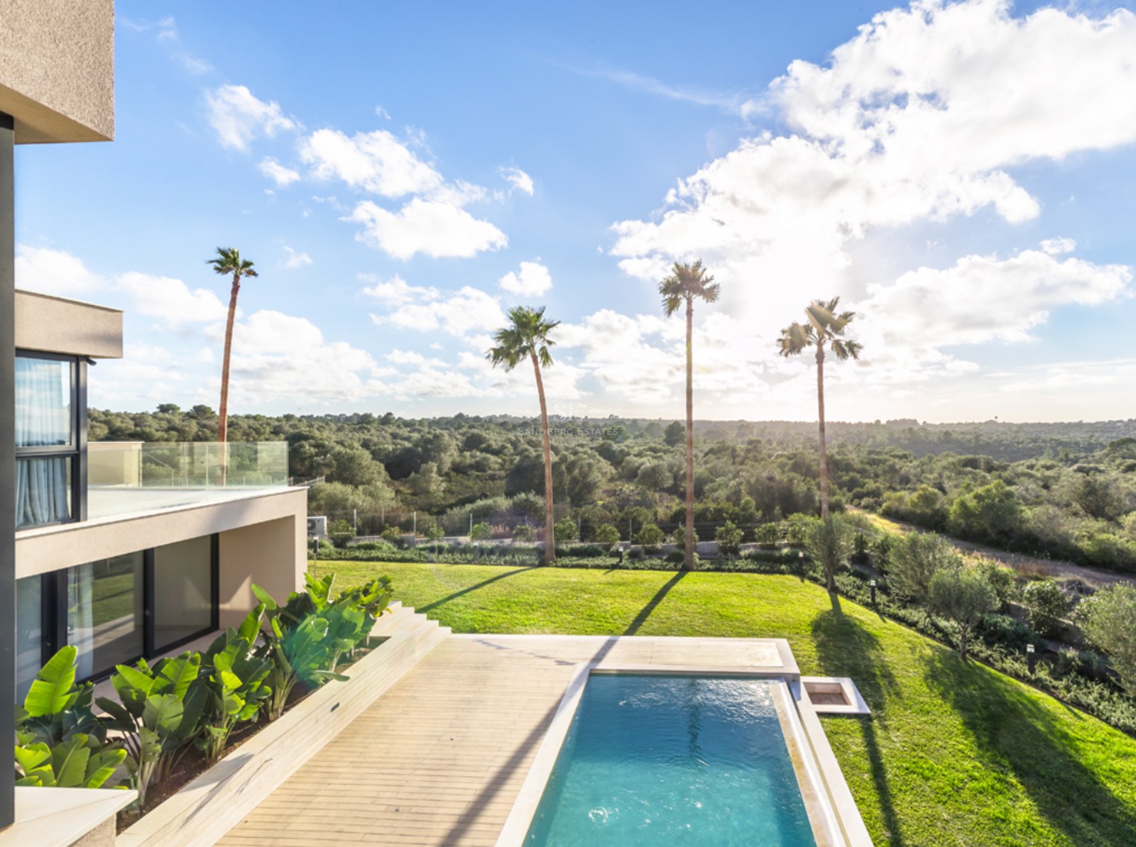Modern villa med en blå pool, trädäck, palmer och utsikt över ett vidsträckt grönt landskap i solljus.