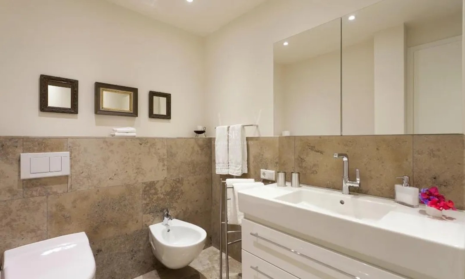 Modern bathroom featuring beige stone wall tiles, a white vanity unit, bidet, and a large wall-mounted mirror.