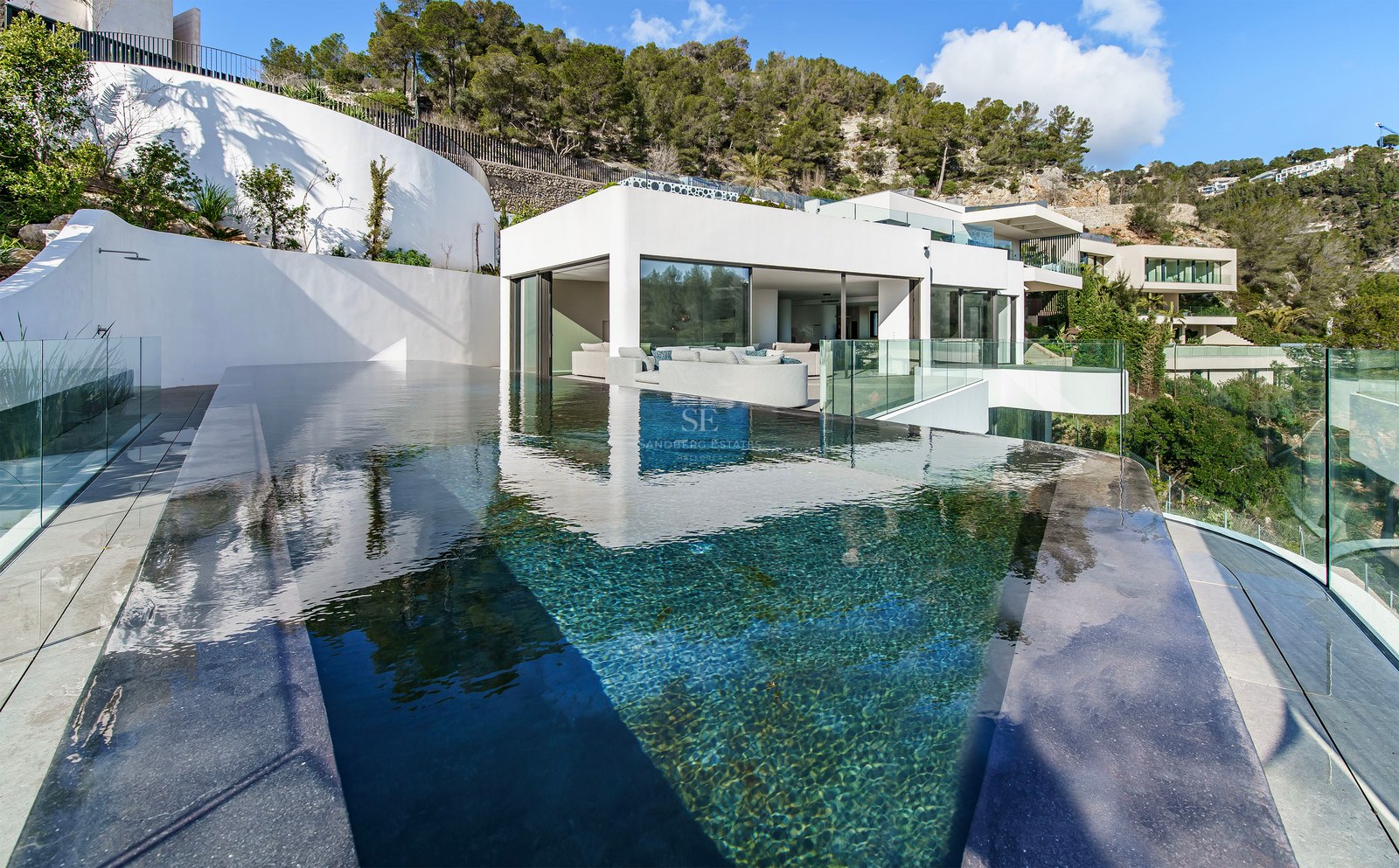 Piscine à débordement moderne aux eaux cristallines surplombant une villa blanche minimaliste nichée dans une colline.