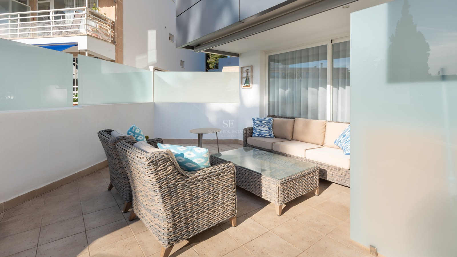 Modern terrace with grey wicker sofas, beige cushions, and blue pillows on a sunlit tiled patio with glass privacy walls.