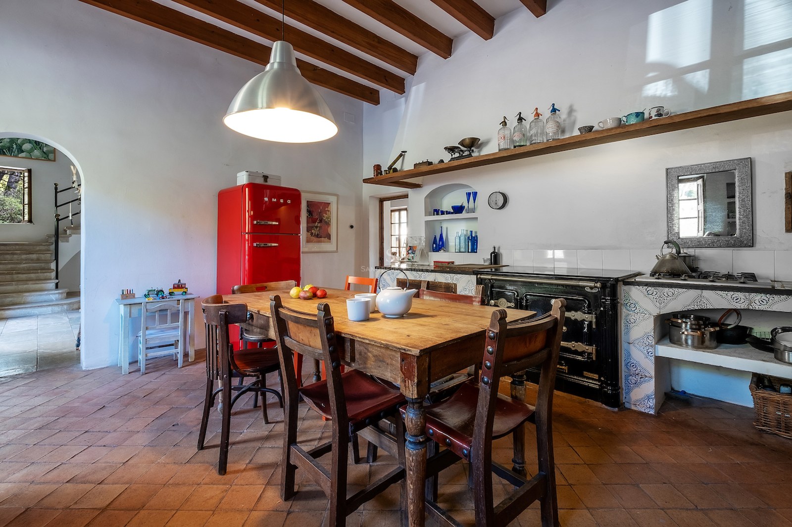 Cuisine traditionnelle avec sols en terre cuite, poutres en bois, table rustique et réfrigérateur rouge vif.