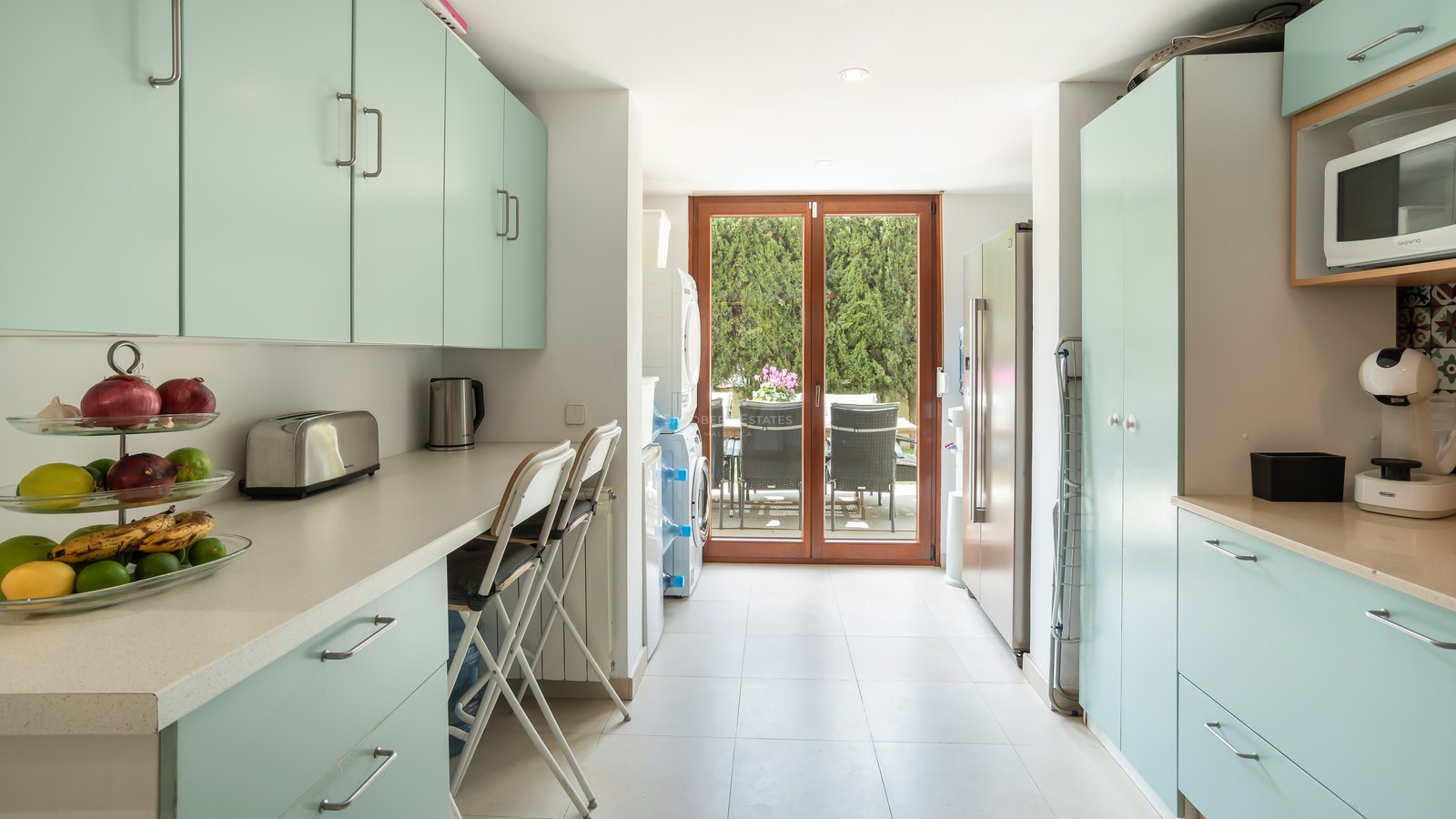 Cocina luminosa con armarios verde menta, encimeras blancas, zona de lavandería y puertas de cristal hacia el patio.