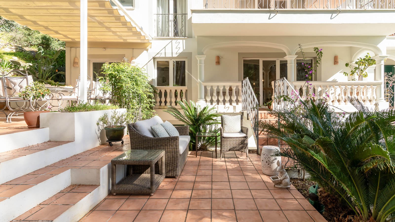Terrasse ensoleillée en terre cuite avec mobilier en osier et plantes devant une façade de villa blanche.
