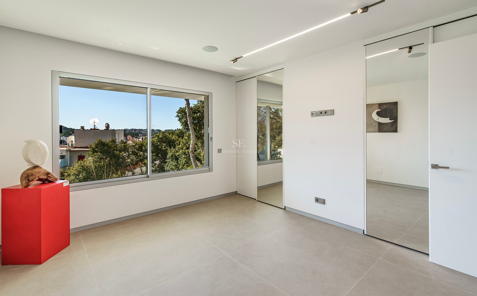 Chambre moderne avec sol gris, placards en miroir et grande fenêtre donnant sur la verdure.