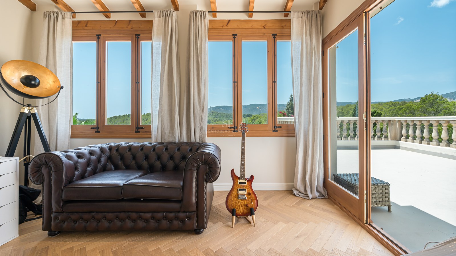 Living room with brown leather Chesterfield sofa, oak herringbone flooring, and large windows overlooking hills.