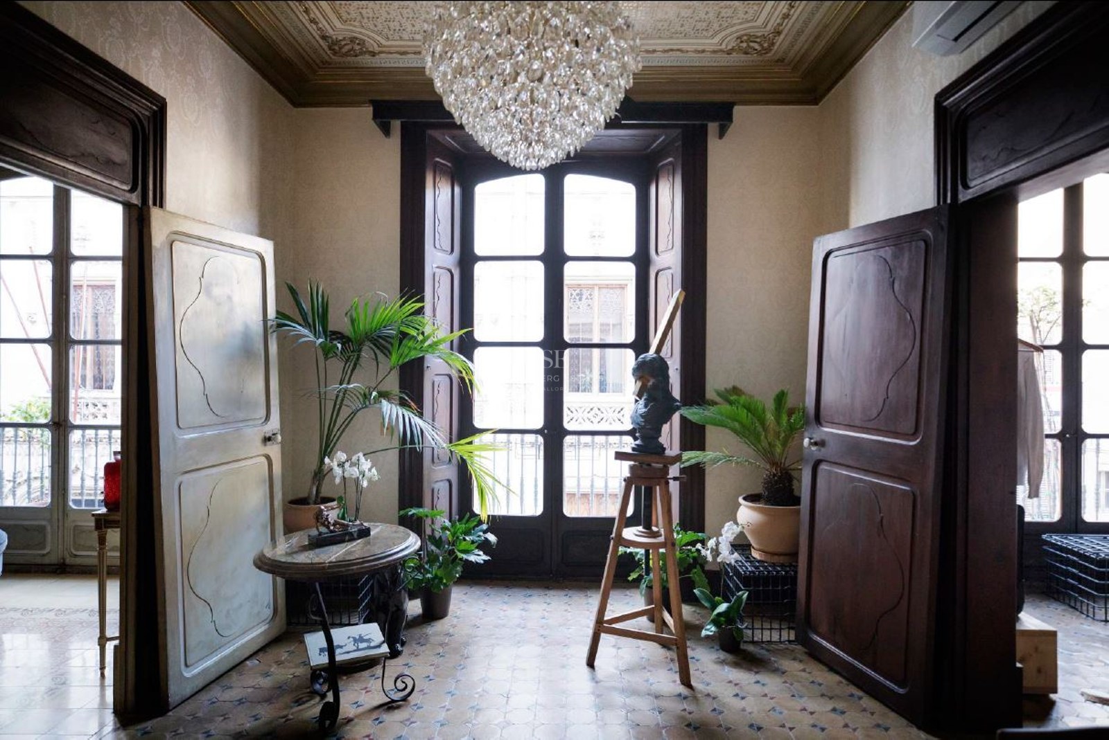 Grand entrance with tall wooden doors, mosaic floors, a crystal chandelier, and indoor plants by a balcony window.