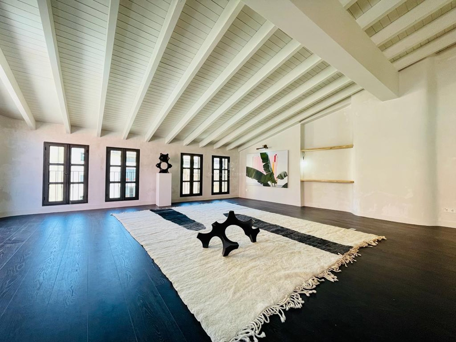 Grande pièce avec parquet foncé, plafonds à poutres blanches et un grand tapis texturé avec sculpture moderne.