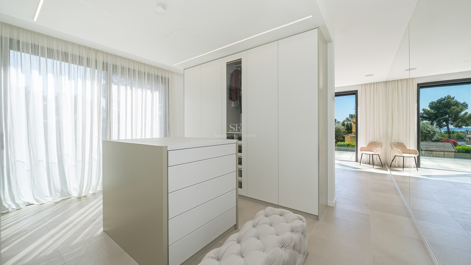 Dressing spacieux avec placards blancs, îlot central, pouf capitonné et vue sur mer par les grandes baies vitrées.