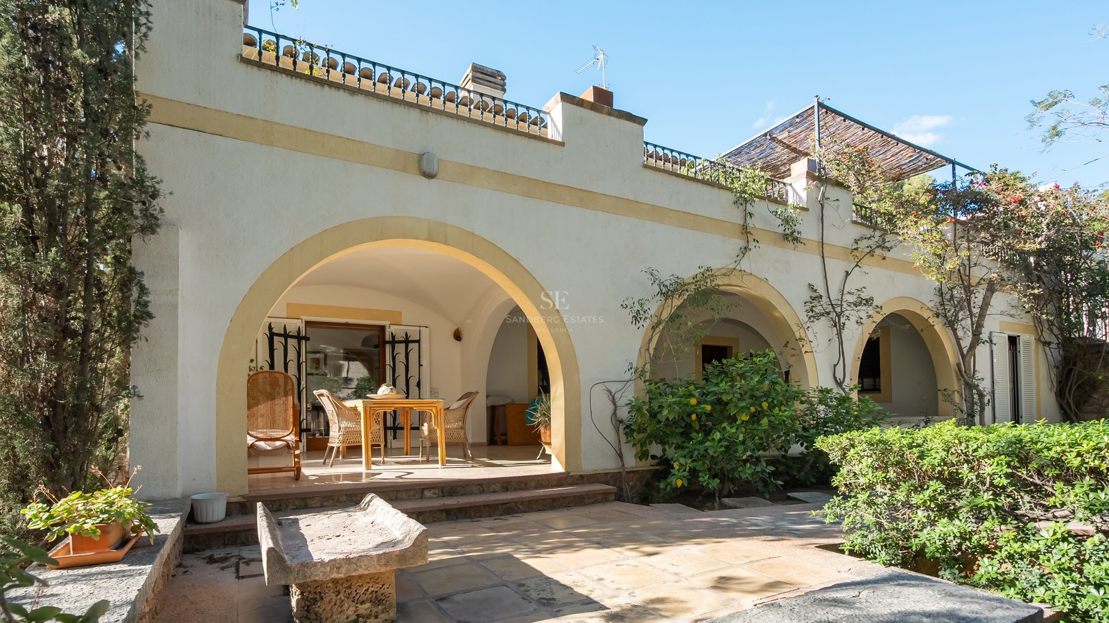 Fachada de villa blanca con grandes arcos, zona de comedor a la sombra, banco de piedra y vegetación mediterránea.