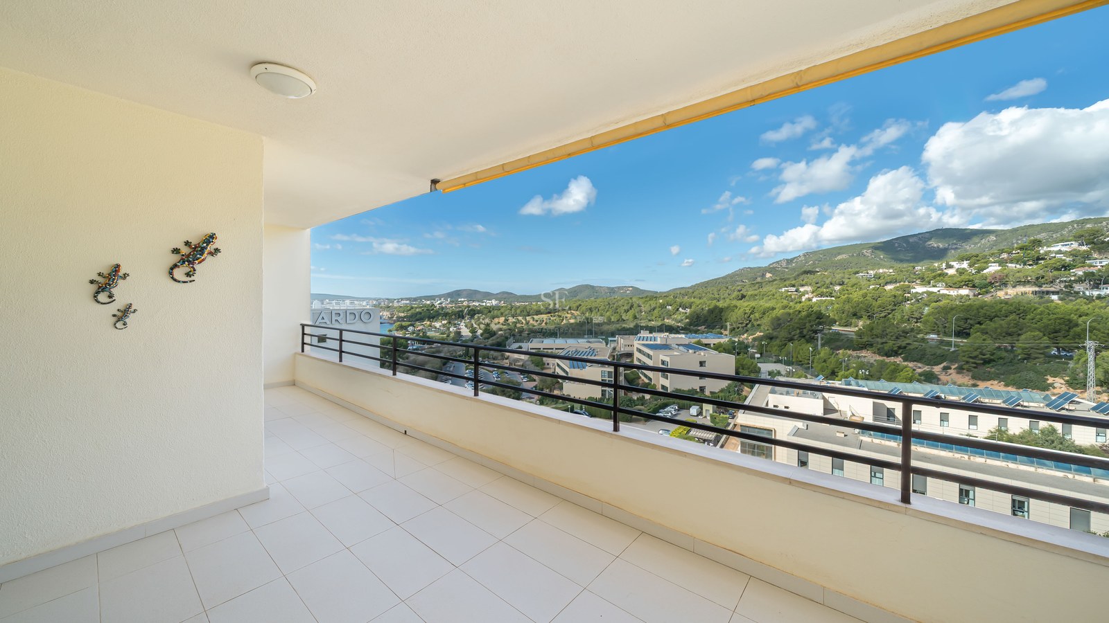 Grand balcon en carrelage blanc avec rambarde noire offrant une vue sur des collines verdoyantes et un ciel bleu.