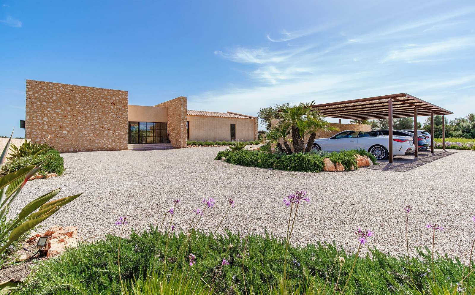 Villa moderne avec murs en pierre naturelle, large allée de gravier et abri voiture sous un ciel bleu pur.