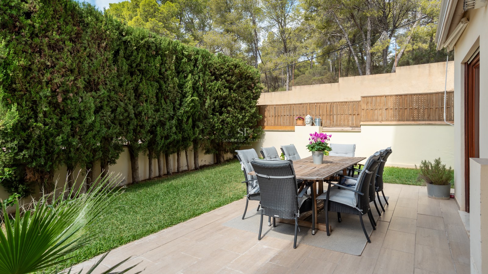 Mesa de madera y ocho sillas en terraza rodeada de altos setos de ciprés y jardín.