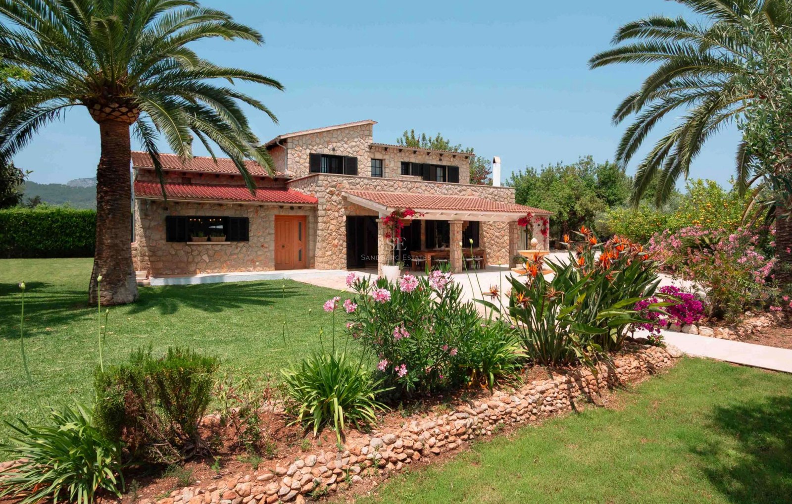 Two-story natural stone villa with terracotta roofs, lush lawn, palm trees, and exotic flowers under a clear blue sky.