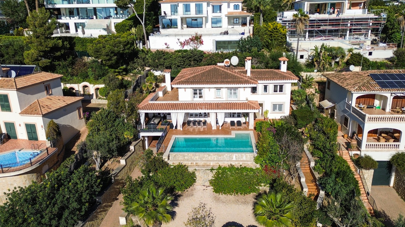 Vue aérienne d'une villa blanche à toit de tuiles avec piscine turquoise et jardins luxuriants.