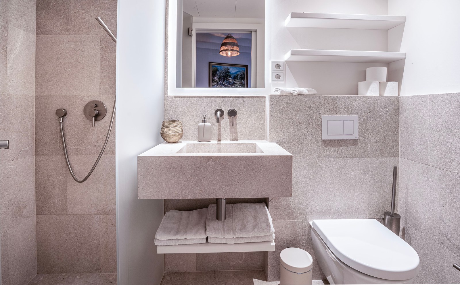 Minimalist bathroom with stone walls, integrated sink, wall-hung toilet, and chrome hand shower.
