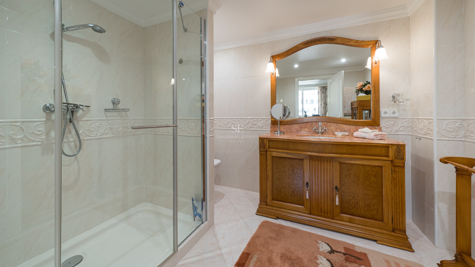 A classic bathroom featuring a glass-enclosed shower, cream marble wall tiles, and a large wooden vanity with a mirror.