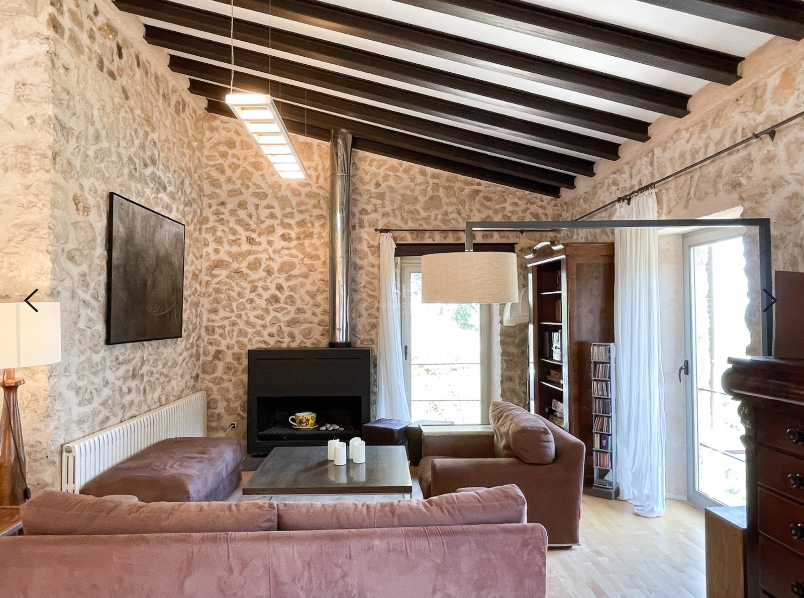 Salón con paredes de piedra natural, techo de vigas de madera oscura, chimenea moderna y ventanales con luz natural.