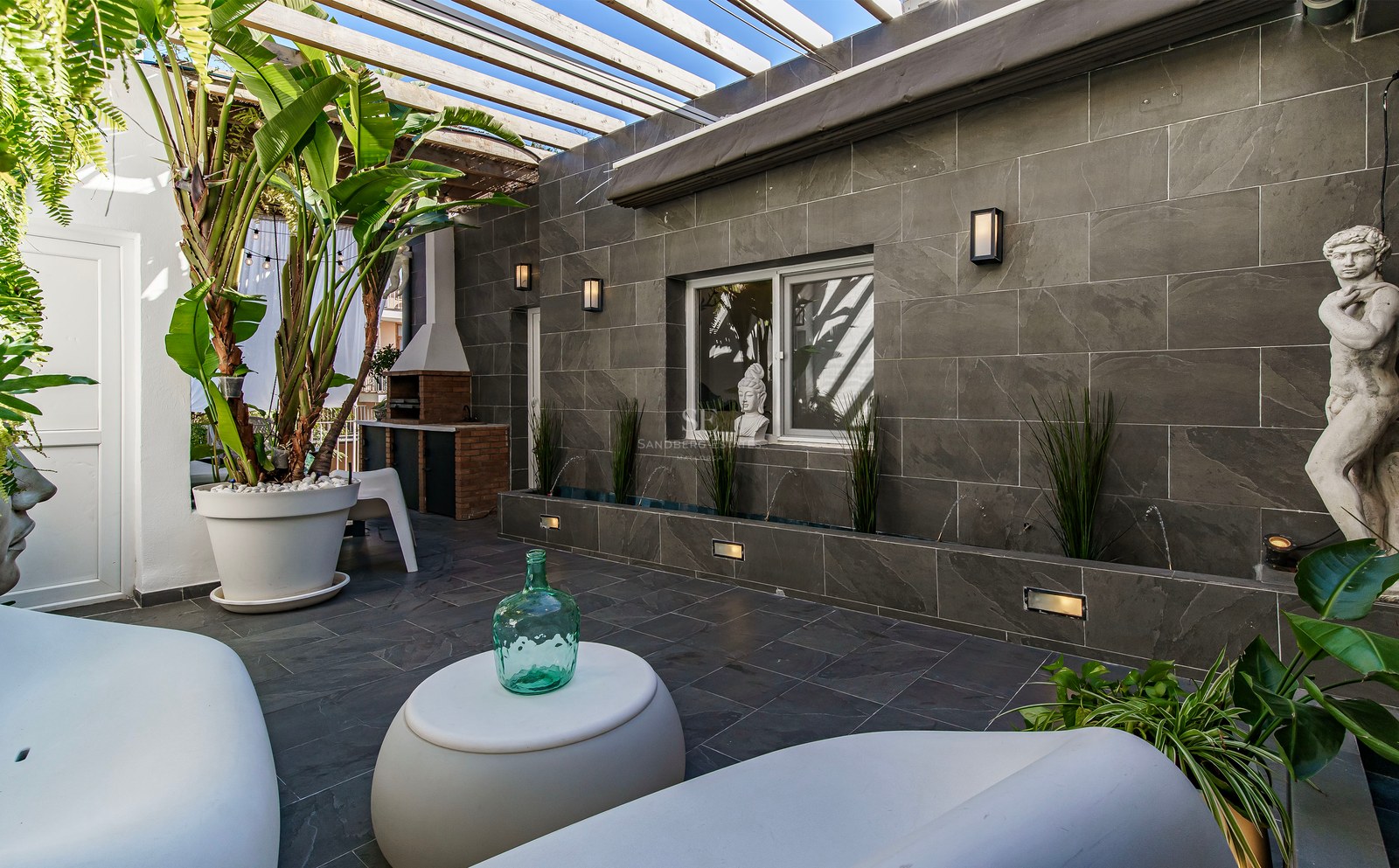Patio de luxe avec murs en pierre grise, mobilier moderne blanc, pergola en bois et plantes tropicales.