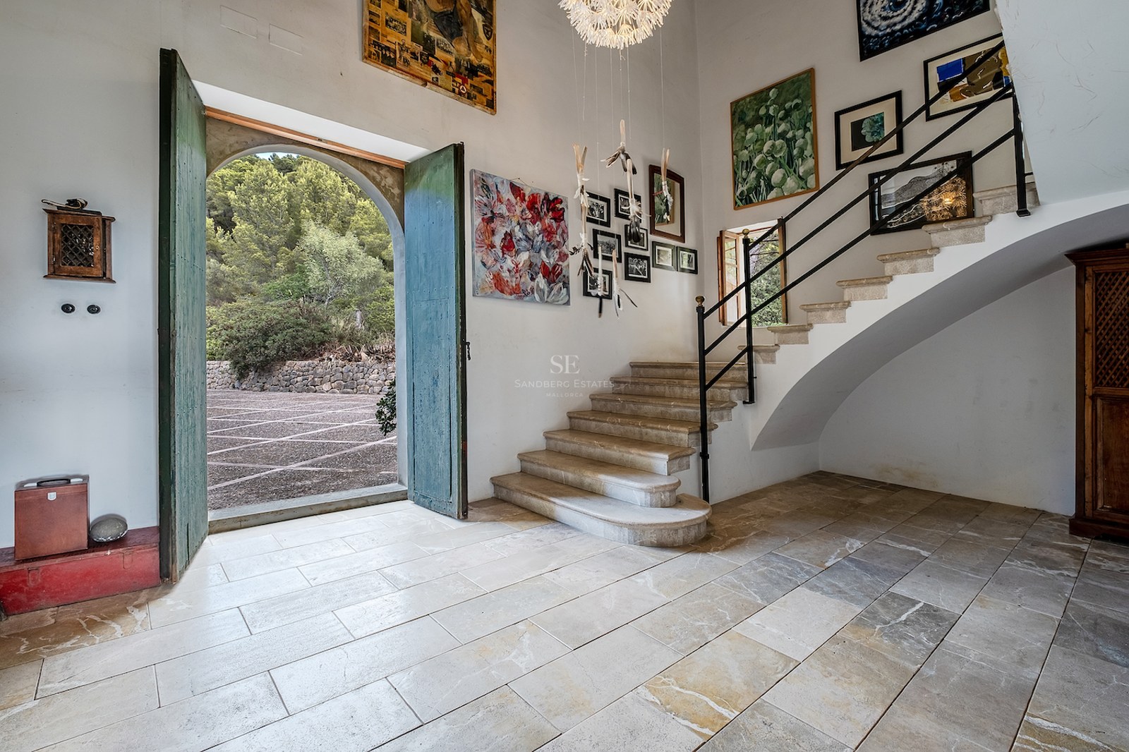 Vaste hall d'entrée avec sols en pierre, hauts plafonds, escalier en pierre et grandes portes vertes ouvertes.