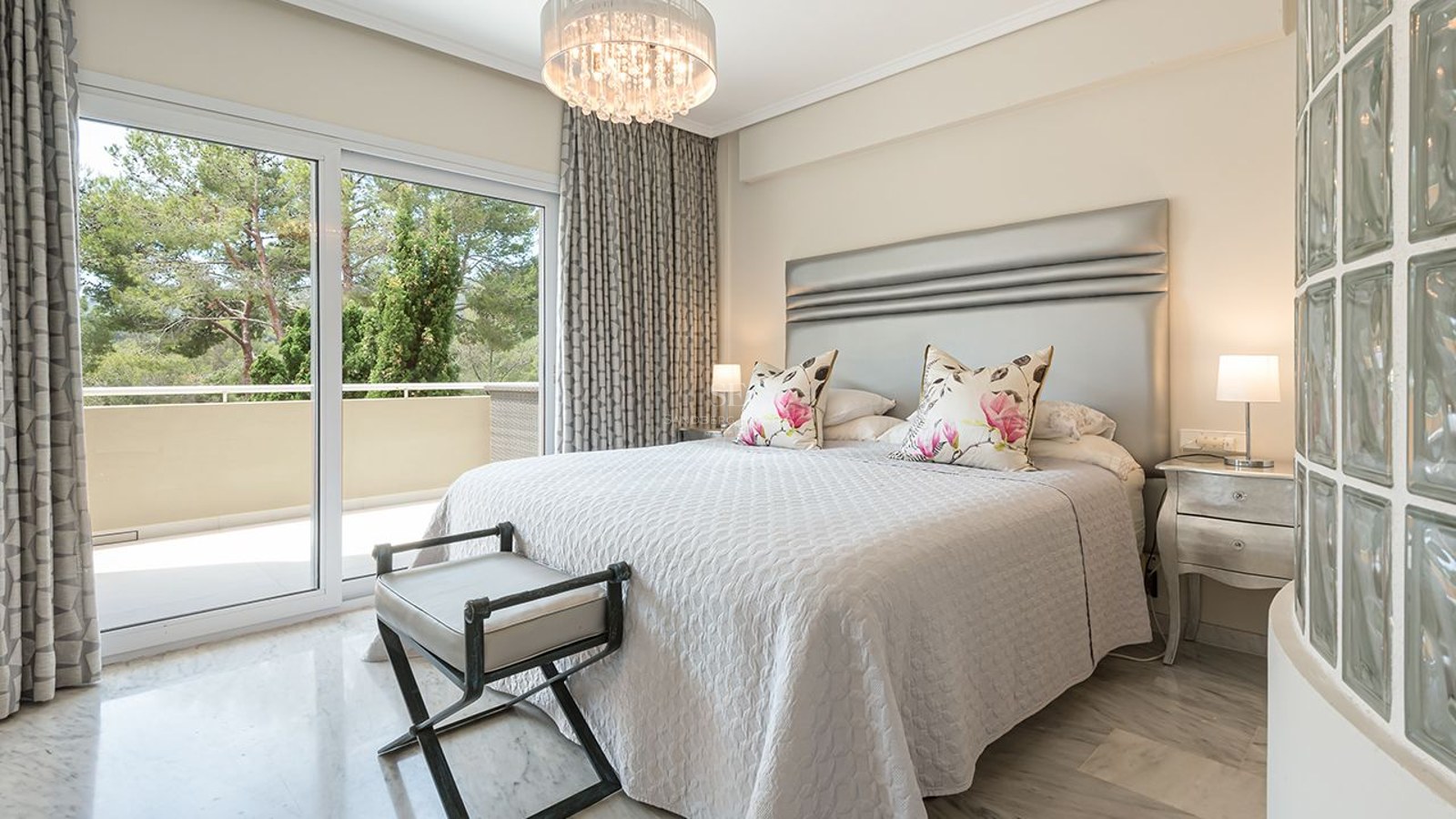 Chambre principale lumineuse avec sol en marbre, tête de lit argentée et accès terrasse vue sur les arbres.