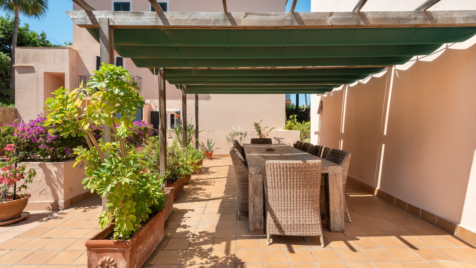 Matplats utomhus på en terrass med terrakottaplattor, träpergola, grön markis och korgstolar.