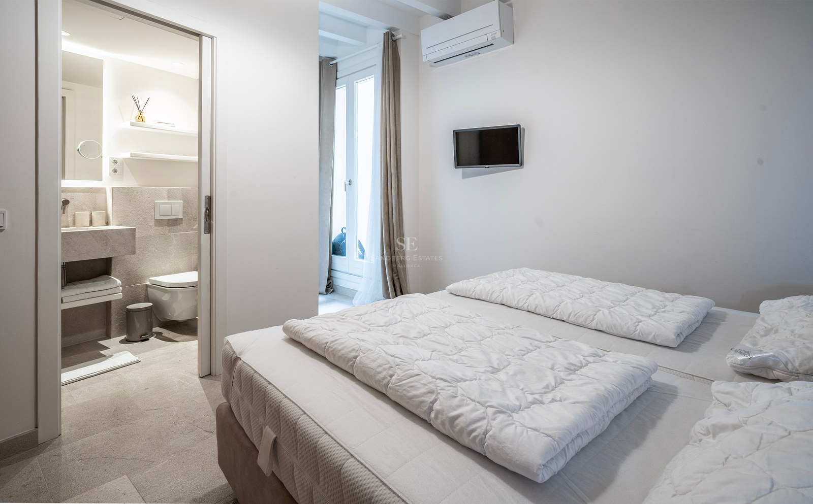Bright modern bedroom with white bedding and open door to a stone-tiled ensuite bathroom.