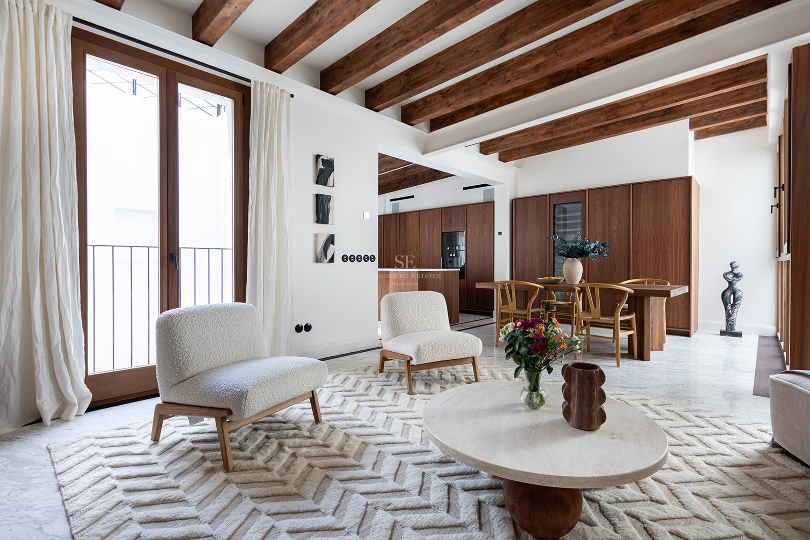 Modern living room with dark wood ceiling beams, white bouclé chairs, marble floor, and integrated dining area.