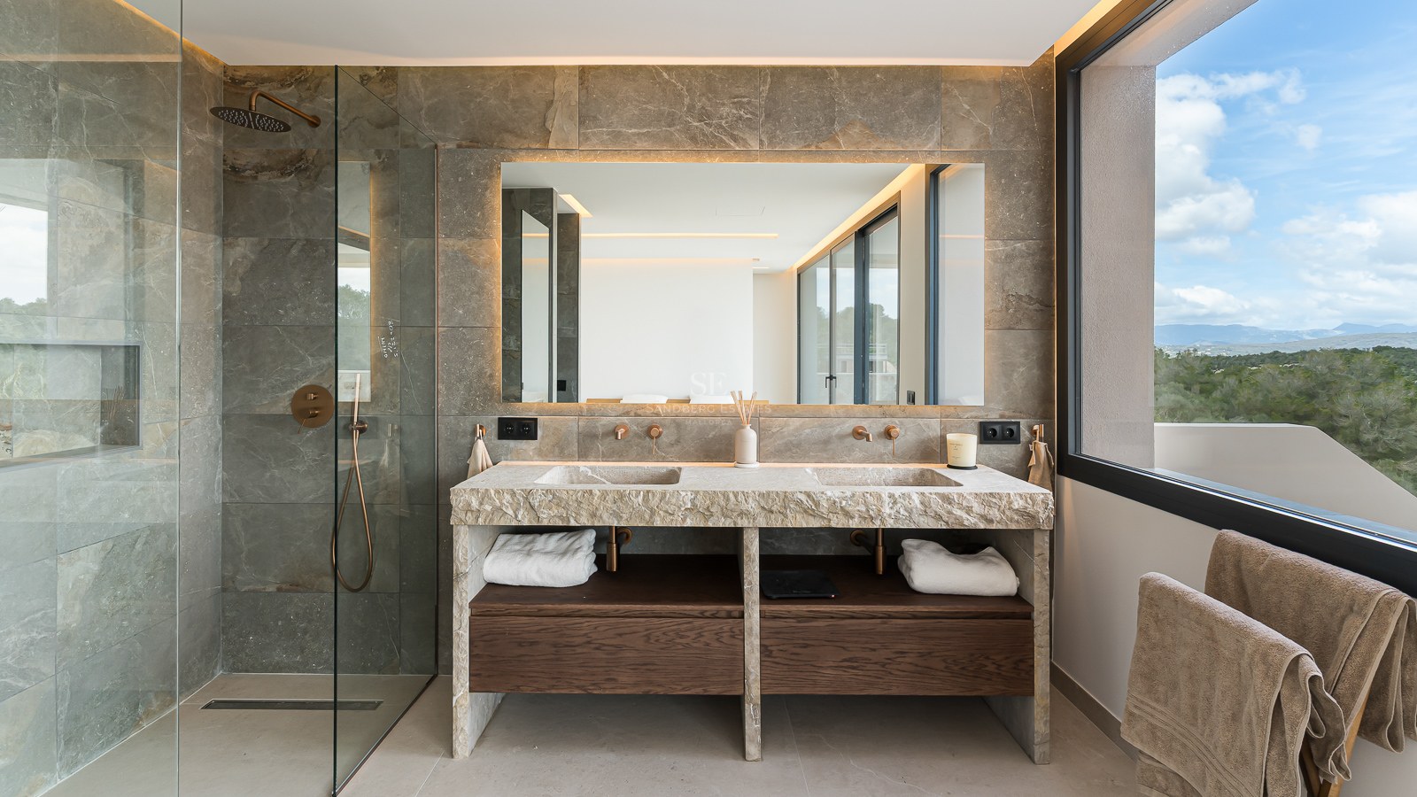 Luxury bathroom featuring a double natural stone vanity, large backlit mirror, glass shower, and dark wood cabinetry.