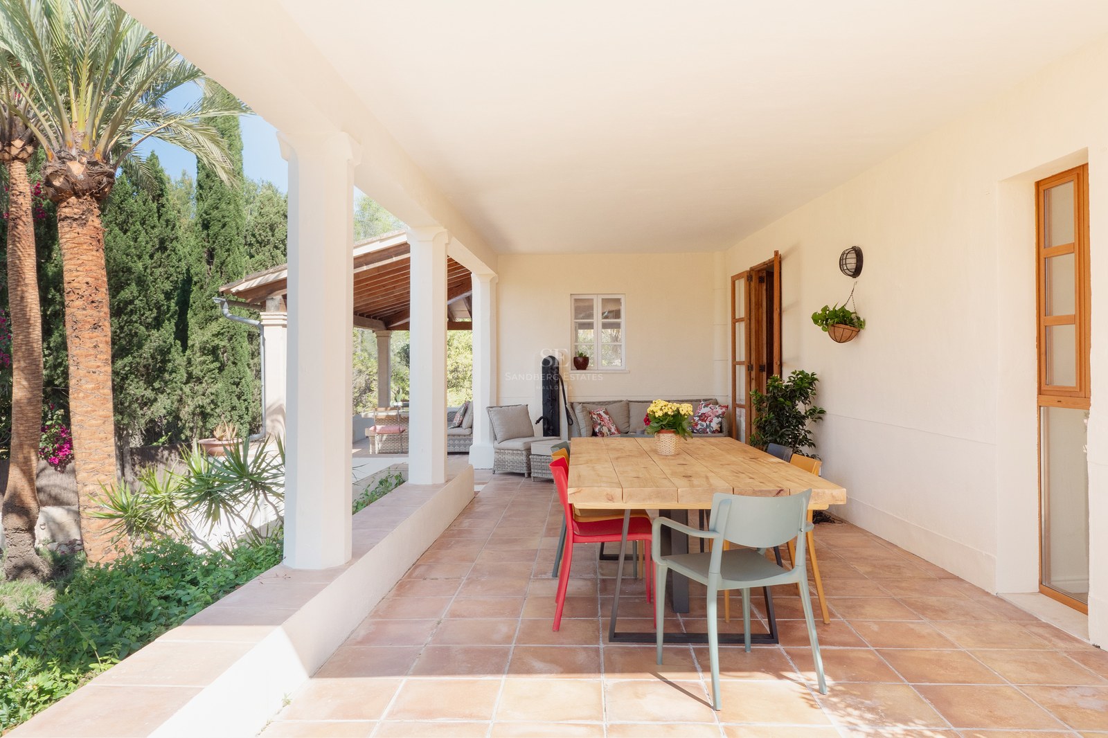Covered terrace with terracotta floors, a large wooden dining table, and view of lush garden with palm trees.
