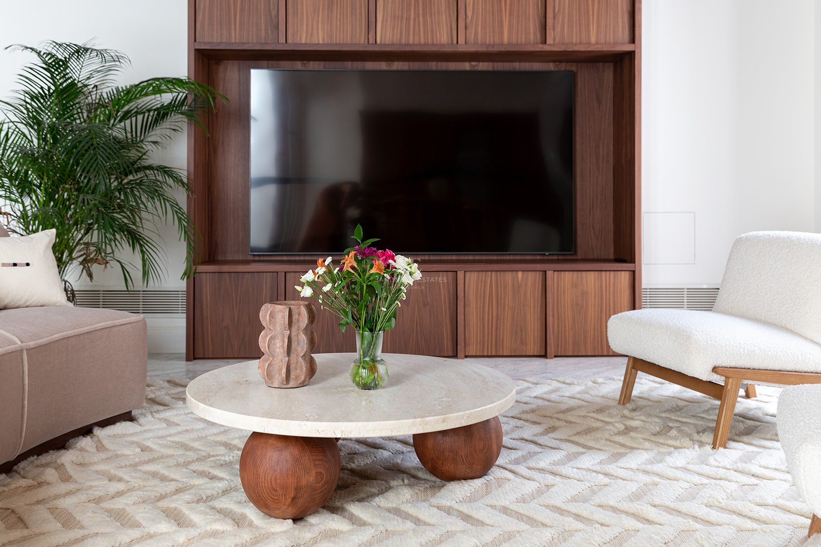 Contemporary living room featuring a walnut TV unit, round travertine coffee table, and white bouclé armchair.